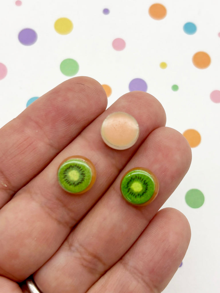 A hand holding two small, round earrings shaped like kiwi fruit against a background of colorful polka dots.