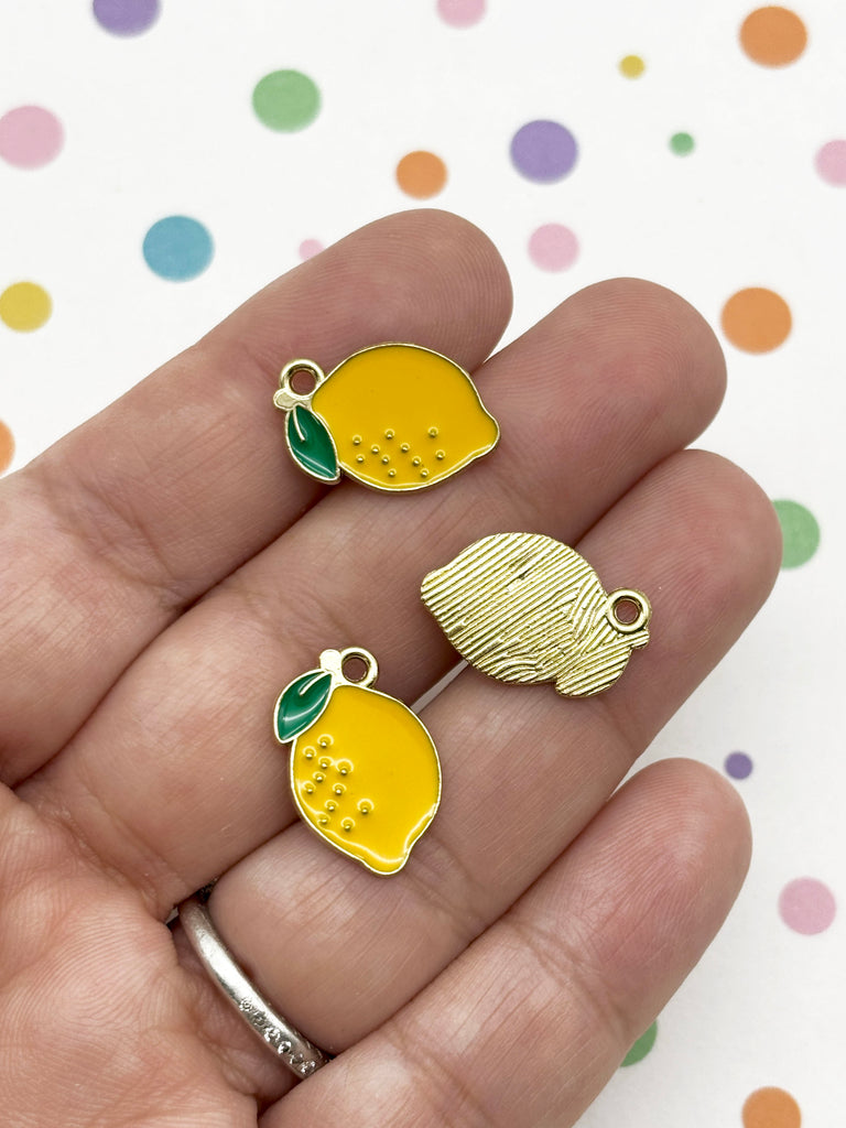 A hand holding three small, yellow enamel charms shaped like lemons against a background with colorful polka dots.