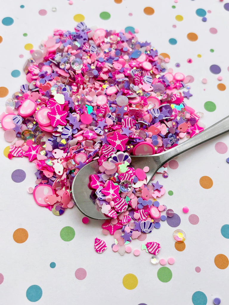 a pile of colorful confetti, including pink, purple, and white pieces, scattered on a white background with polka dots. A spoon is visible, suggesting the confetti was recently used or is about to be used.