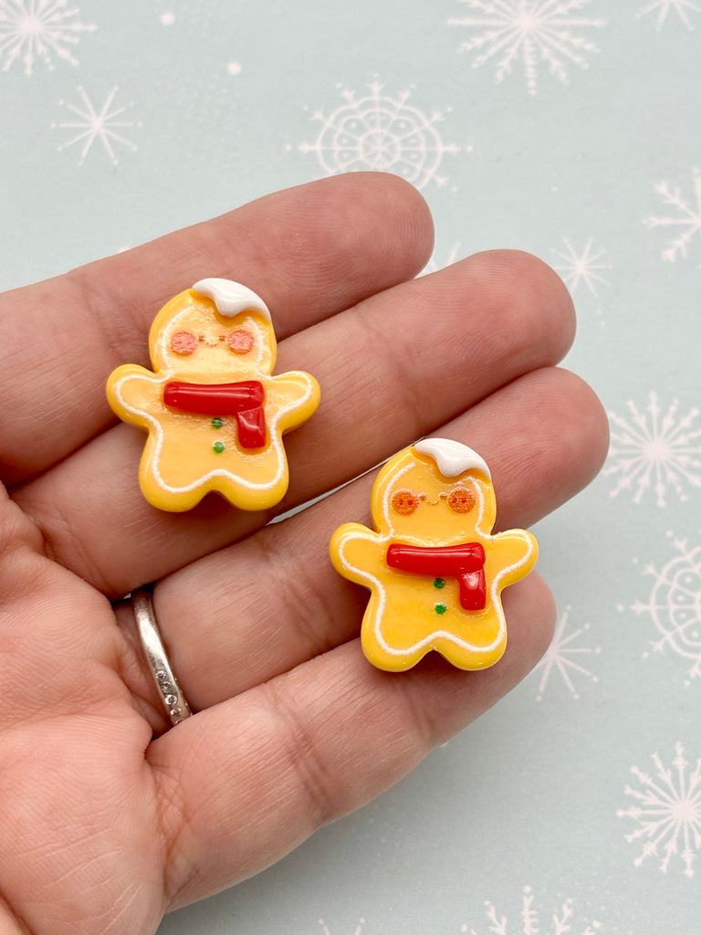 A hand holding two gingerbread man-shaped earrings against a snowy background.