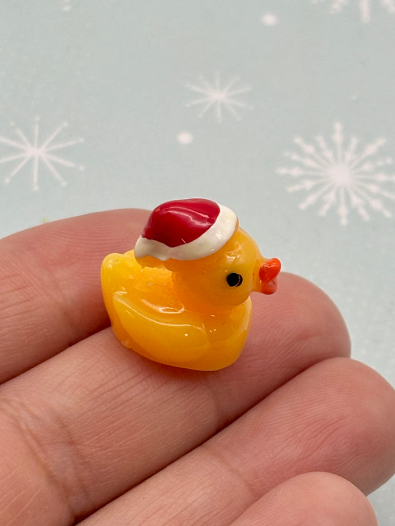 A small yellow rubber duck wearing a red Santa hat is being held in a person's hand against a background of snowflakes.