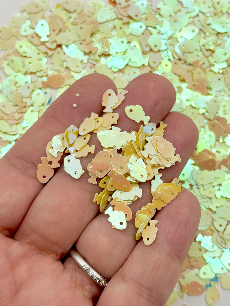A hand holding a collection of small, colorful fish-shaped sequins against a background of similar fish-shaped sequins.