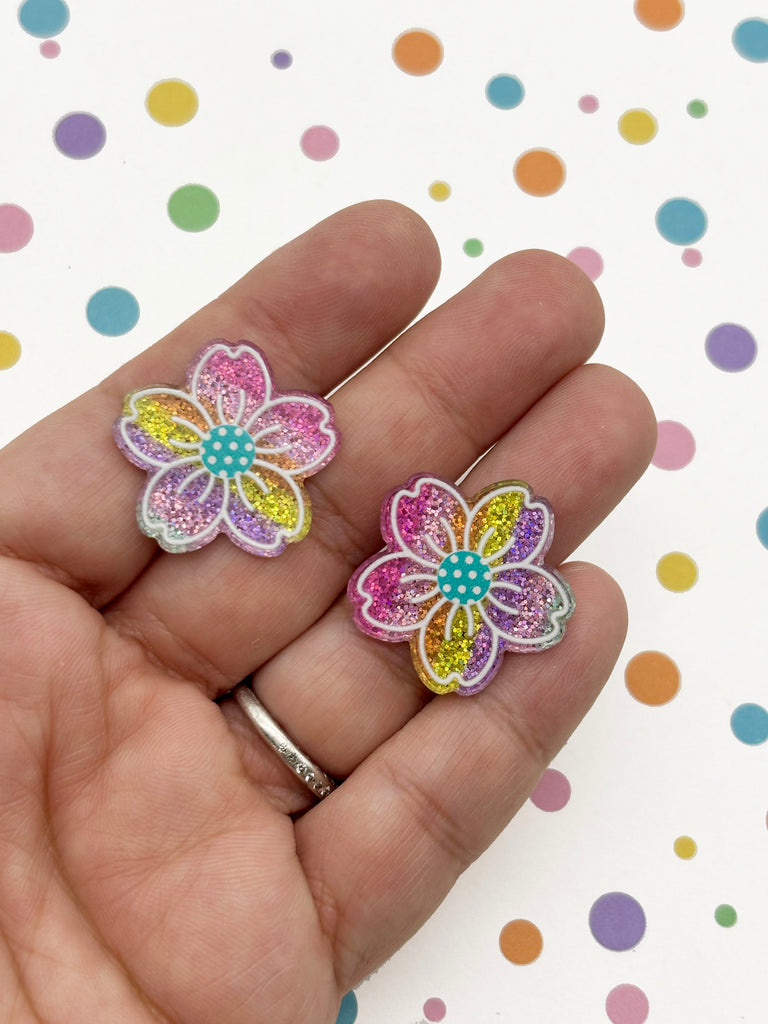 A hand holding two colorful, glittery flower-shaped earrings against a background of multicolored polka dots.