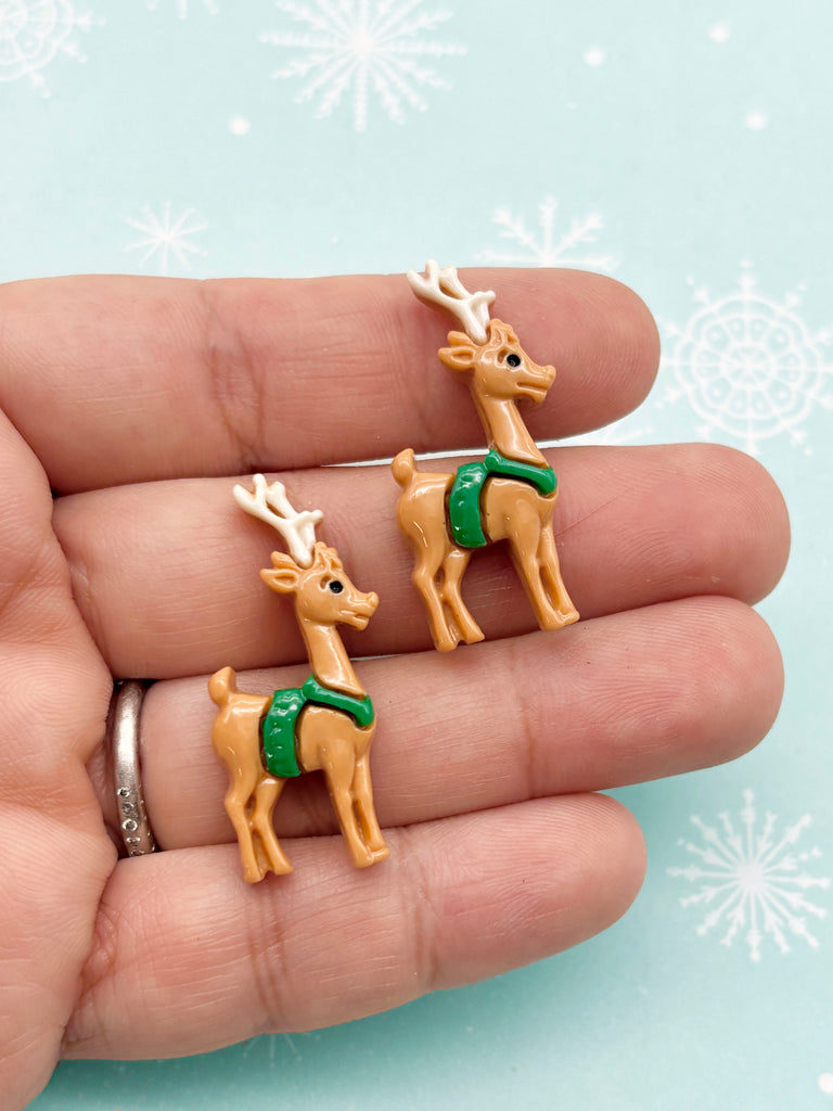 A hand holding two small, brown reindeer figurines with green ribbons on their necks against a light blue background with white snowflakes.