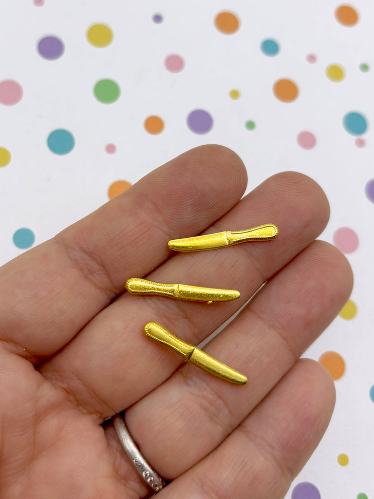 A hand holding three small gold-colored knife clips against a background with colorful polka dots.