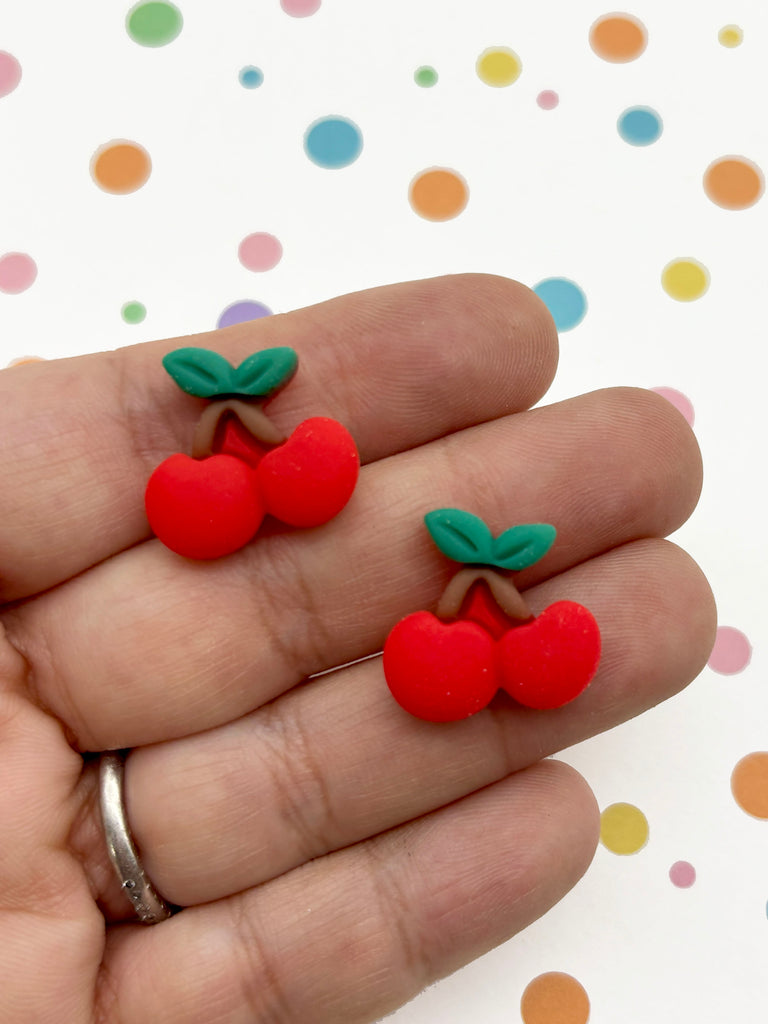 A hand holding two red plastic cherries against a background with colorful polka dots.