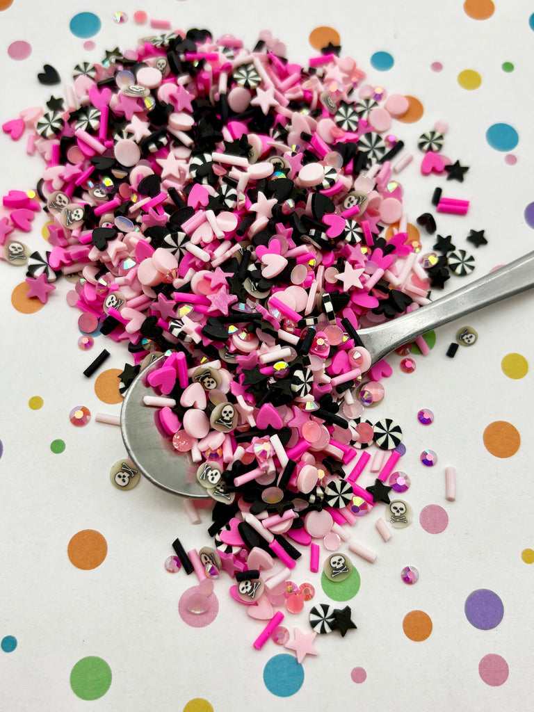a pile of colorful sprinkles, including pink, black, and white, scattered on a white surface with polka dots. A spoon is visible among the sprinkles, suggesting the sprinkles have been recently used or are being used for a creative purpose.