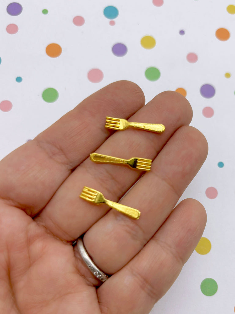 A hand holding three small gold-colored forks against a background with colorful polka dots.