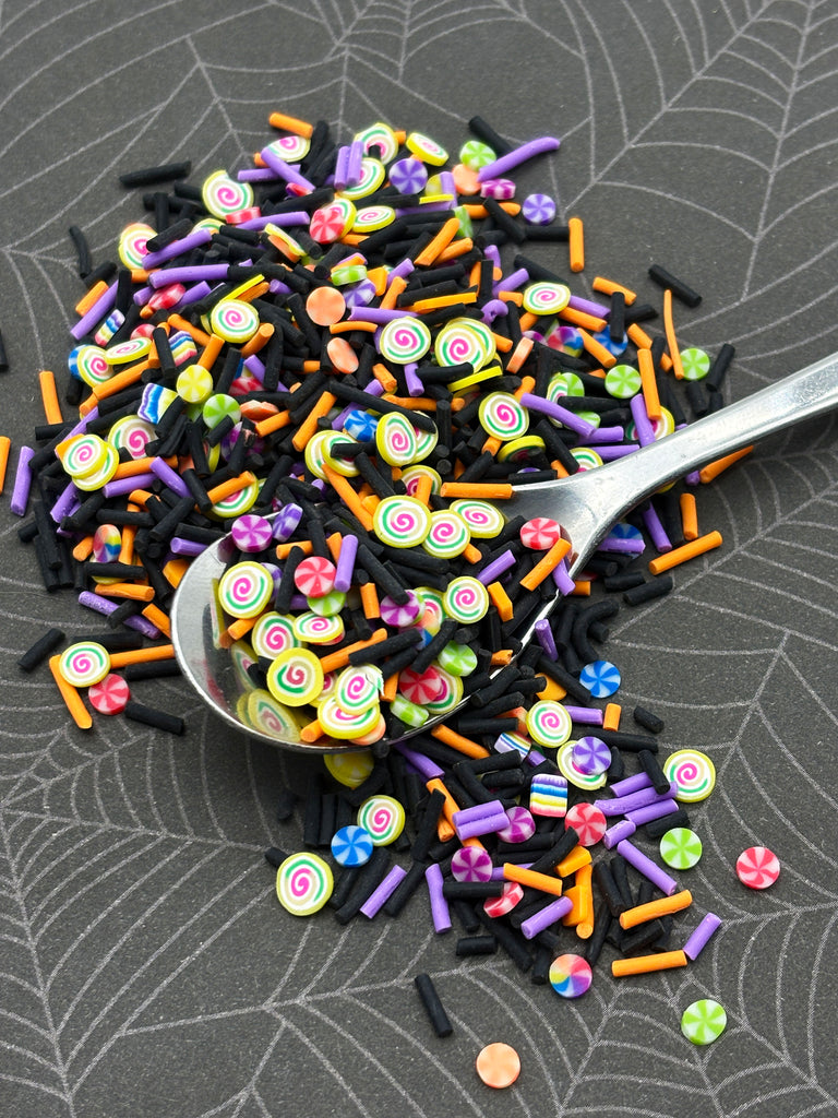 A silver spoon is scooping up a colorful assortment of candy sprinkles, including black, orange, purple, and yellow, on a gray surface.