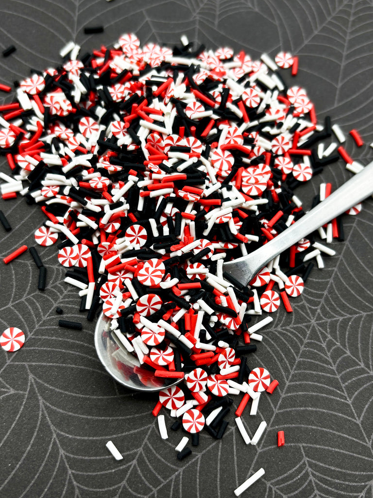 a pile of colorful sprinkles, including red, white, and black, scattered on a dark surface. A spoon is visible, suggesting the sprinkles were recently used or are about to be used.