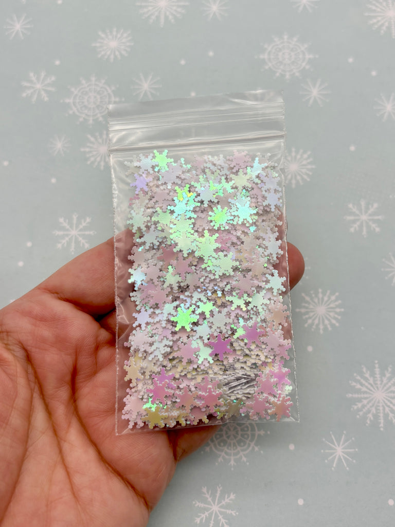 A hand is holding a clear plastic bag filled with colorful glittery star-shaped objects against a snowy background.