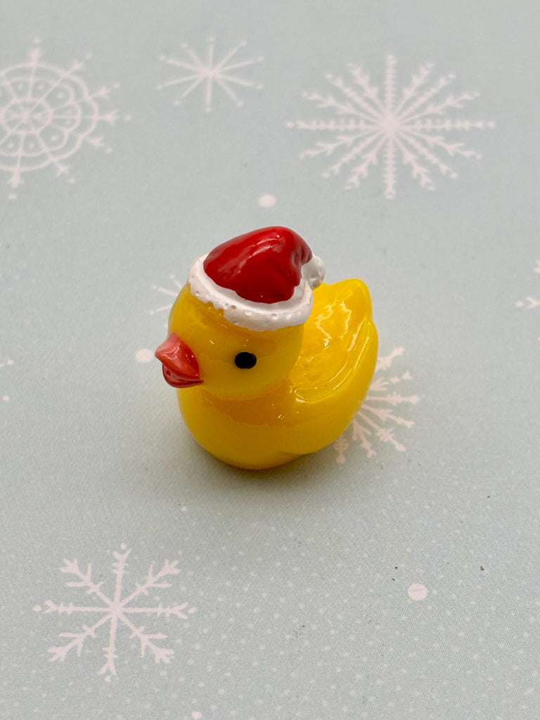 A yellow rubber duck wearing a red Santa hat is sitting on a light blue background with white snowflake designs.