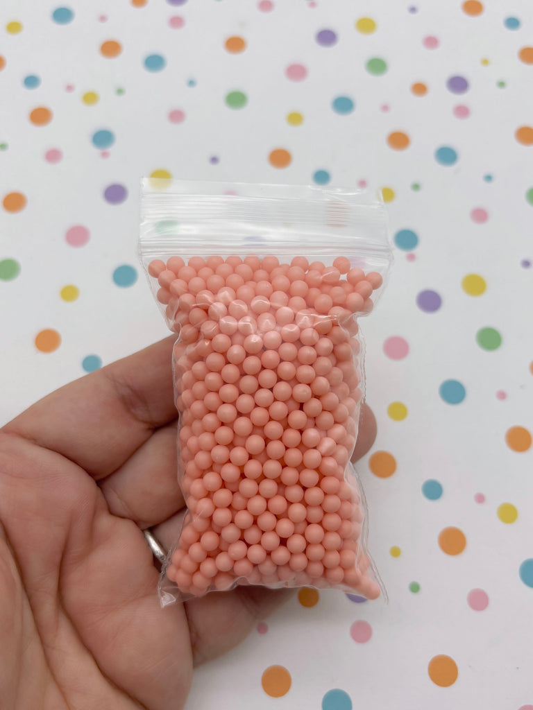 A hand is holding a small bag filled with small, round, pink beads against a background with colorful polka dots.