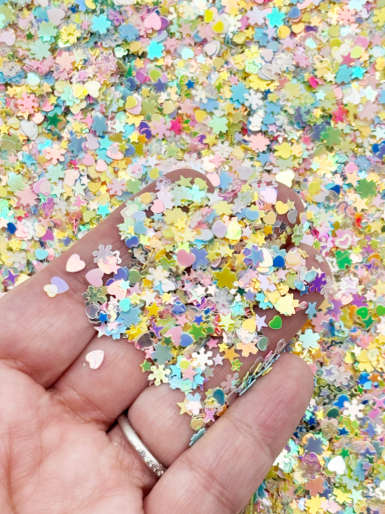 A hand holding a pile of multicolored glitter, including hearts, stars, and flowers, against a background of similar multicolored glitter.