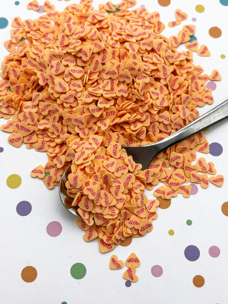 a pile of orange heart-shaped sprinkles on a white surface with colorful polka dots. A spoon is visible, and the sprinkles are scattered around it.