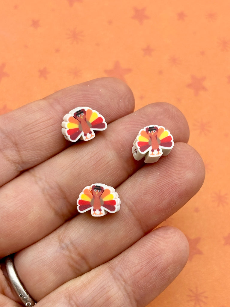 A hand holding three small, colorful beads shaped like turkeys against an orange background with stars.