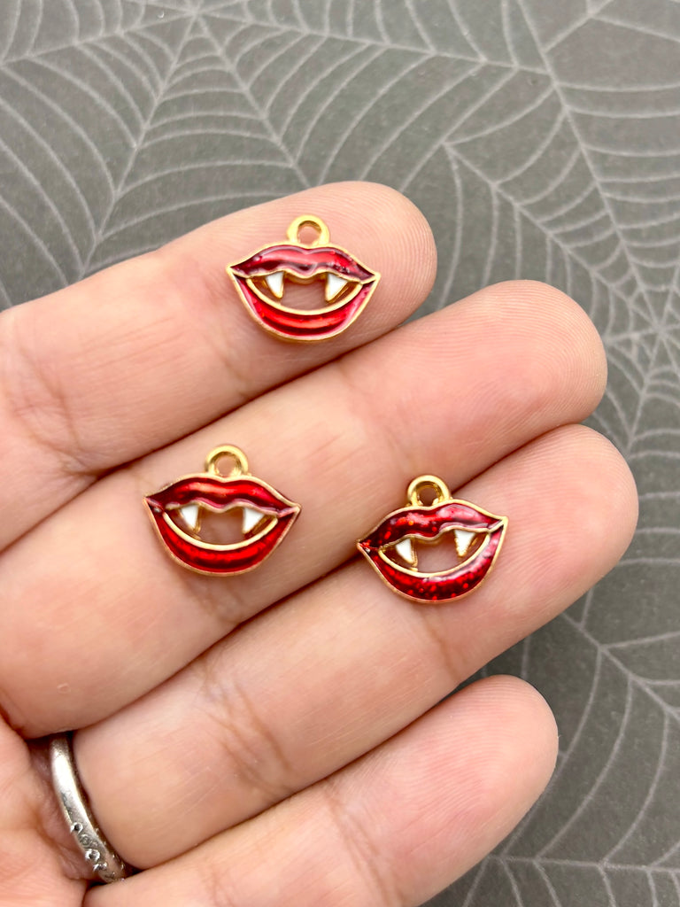 A hand holding three small red and gold charms shaped like teeth, resembling a vampire or monster's mouth.