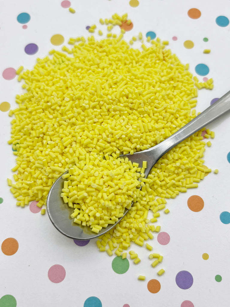 a pile of yellow sprinkles on a white surface with colorful polka dots. A spoon is resting on top of the sprinkles, and the background is dotted with various colors.