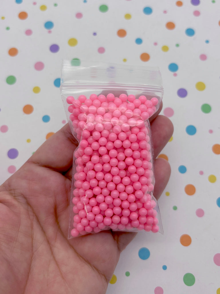 A hand is holding a clear plastic bag filled with small, round, pink beads against a background with colorful polka dots.