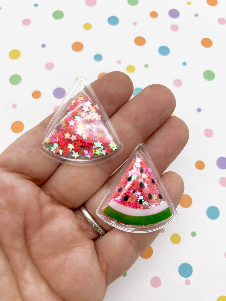 A hand holding two clear plastic objects shaped like watermelon slices, decorated with glitter and stars.