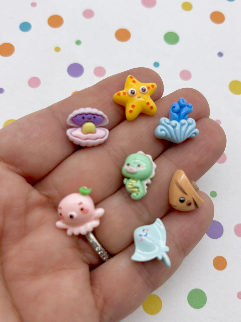 A hand holding a collection of colorful, whimsical sea animal-themed charms, including a starfish, seashell, jellyfish, and other playful creatures.