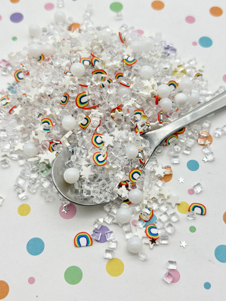 a spoon filled with rainbow-colored candy pieces, including white and clear beads, surrounded by scattered candy pieces on a white background with colorful polka dots.