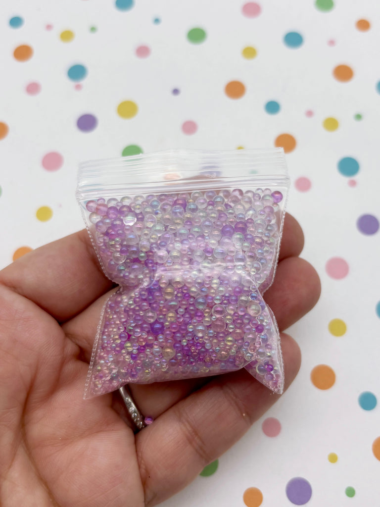 A hand is holding a small bag filled with small, clear beads in various colors, including purple and pink, against a background with colorful polka dots.