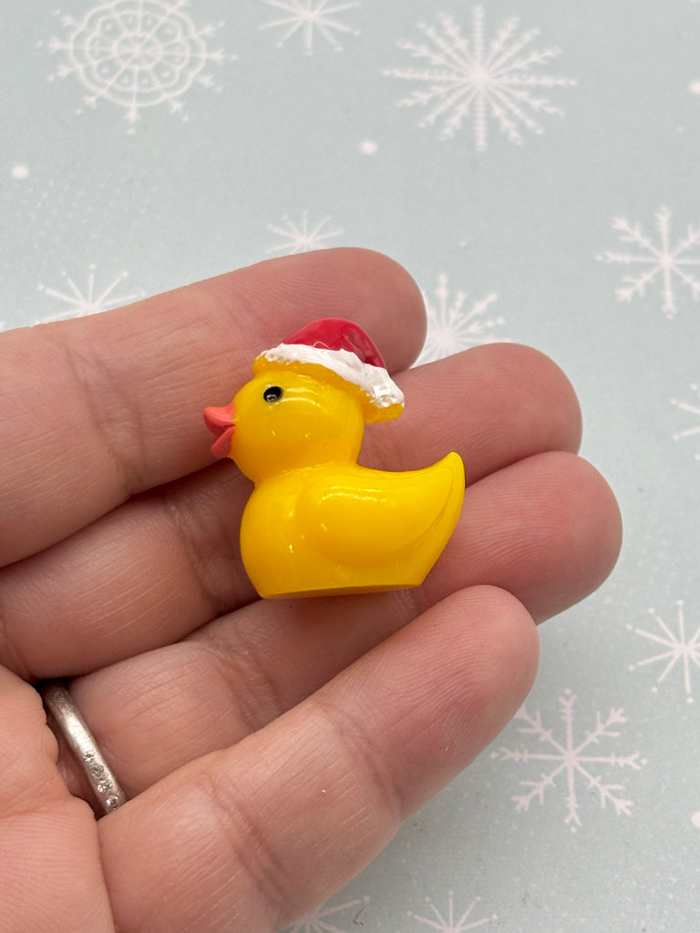 A small yellow rubber duck wearing a red Santa hat is being held in a person's hand against a background of snowflakes.