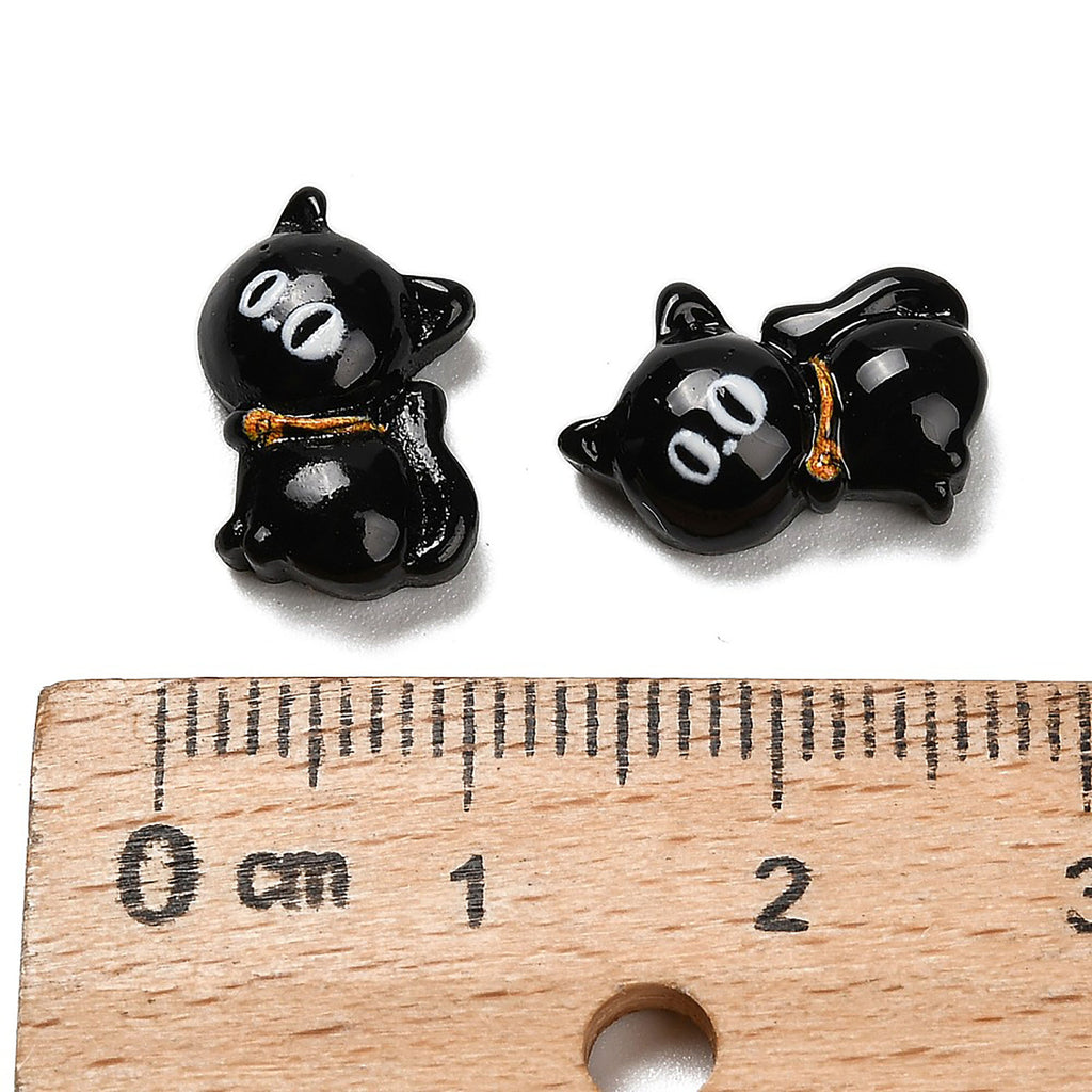 two black cat-shaped charms with white eyes, placed above a ruler for size comparison.